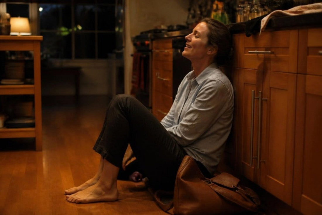 A woman just home from work sitting on the kitchen floor with her shoes off, letting out a long exhale