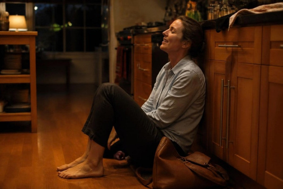 A woman just home from work sitting on the kitchen floor with her shoes off, letting out a long exhale