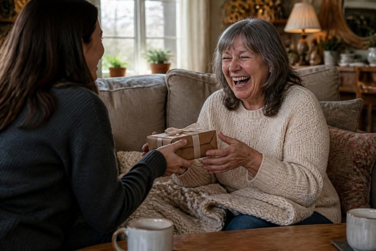 A mom laughing on the sofa as her adult daughter hands her a small wrapped gift