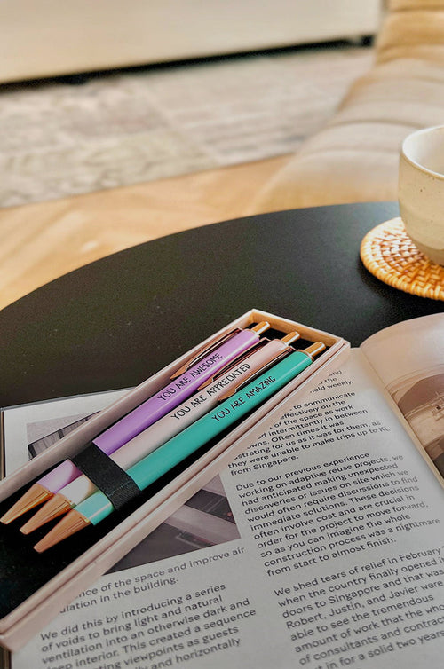 Pastel Mesmos pens held by an elastic band, resting on an open book beside a coffee cup on a dark table.