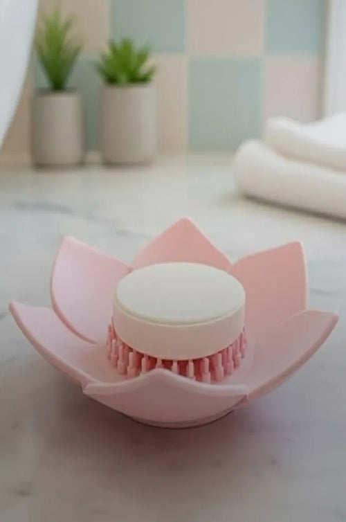 A pink lotus-shaped tray holding a shower steamer on a marble ledge, with plants and folded towels behind.