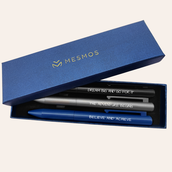 Three dark metal pens in an open navy Mesmos gift box, engraved Dream Big And Go For It, The Adventure Begins and Believe And Achieve.