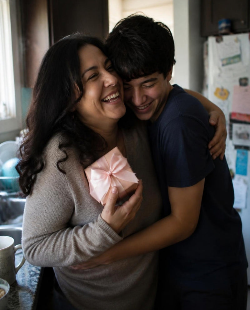 A laughing Hispanic mother hugging her teenage son in the family kitchen, the Mesmos Mom Series box in her hand